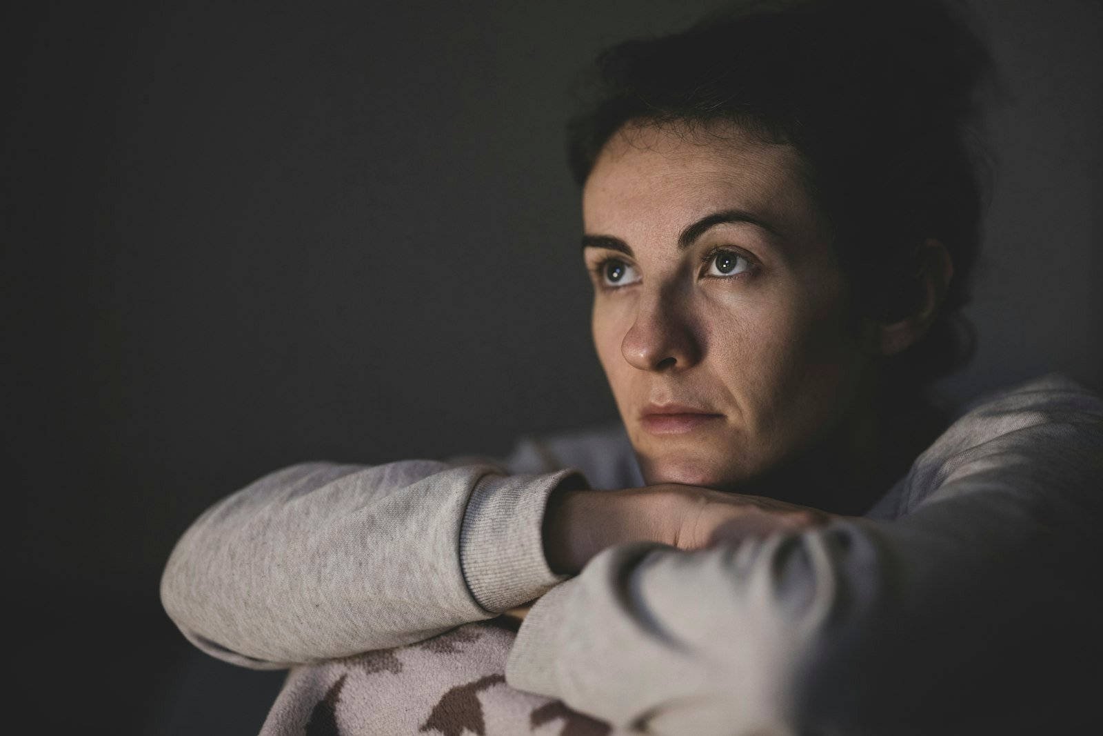 Femme au regard las, le menton posé sur ses bras dans une pièce sombre