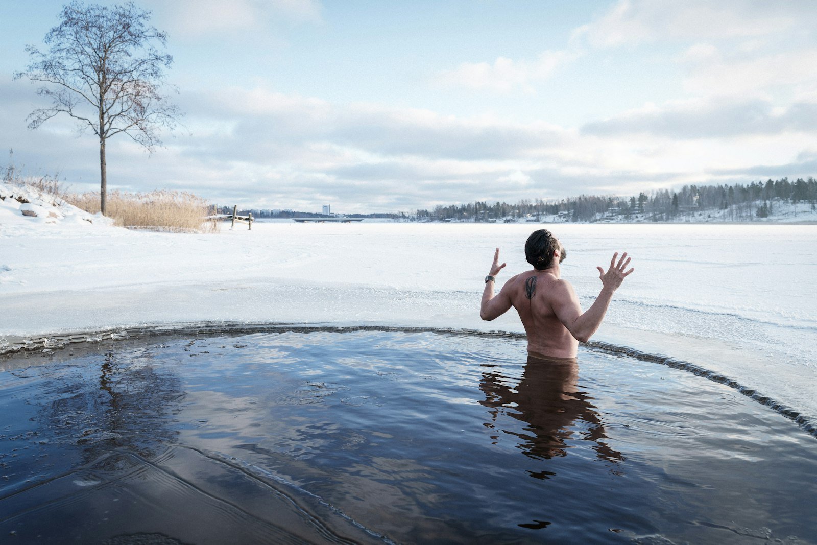Homme immergé dans un trou de glace sur un lac gelé, bras levés, paysage enneigé